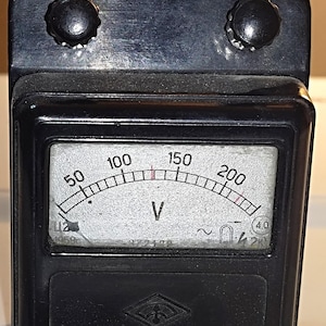 Puede incluir: Un voltímetro negro vintage con un indicador curvo que muestra el voltaje de 50 a 200. La esfera tiene un fondo blanco con marcas negras y una aguja que indica el voltaje. Dos perillas negras están en la parte superior.