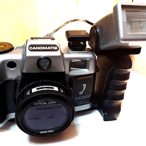 May include: A silver and black Canomatic camera with a flash attachment. The camera has a lens with the text "50mm 1:2 OPTICAL LENS" and "FOCUS FREE". The flash is attached to the top of the camera.