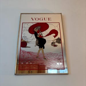 Peut inclure: Un miroir mural rectangulaire orné d'une impression décorative avec le mot VOGUE en haut. L'illustration représente une silhouette en vêtements noirs tenant un grand parapluie rouge, entourée de lignes rouges stylisées de vagues et de nuages.