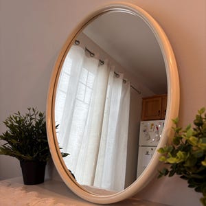 Peut inclure: Un miroir ovale doté d'un cadre lisse de couleur crème. La surface réfléchissante offre une vue claire sur une pièce avec des rideaux blancs, un réfrigérateur et des armoires en bois. Le miroir est positionné verticalement, au design épuré.