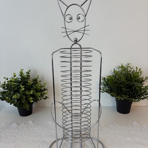 May include: A silver-colored metal cat-shaped paper towel holder featuring a coiled wire body, a circular base, and a stylized head with wire whiskers and ears. The structure is made of thin, polished metal rods forming a vertical cylinder.