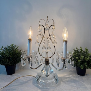 Peut inclure: Une lampe de table candélabre à deux lumières avec une structure en métal doré, des bras incurvés et une base en verre transparent. La lampe est ornée de multiples prismes en cristal transparent en forme de goutte et deux bougies.