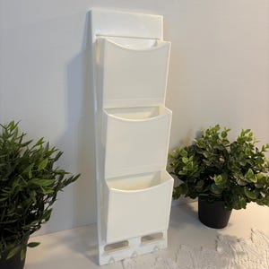 Könnte beinhalten: Ein weißer Wand-Organizer aus Kunststoff mit drei vertikalen Aufbewahrungstaschen mit geschwungenen Oberkanten. Die Einheit verfügt über eine flache Rückwand zur Montage und einen unteren Bereich mit drei kleinen Haken.