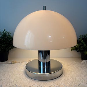 May include: A retro table lamp featuring a cream-coloured, dome-shaped shade and a chrome base. The lamp has a cylindrical chrome stem and a round base. The lamp is on a white surface with a lace doily.