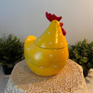 Peut inclure: Un pot en céramique jaune en forme de poule avec des pois blancs. La poule présente une crête et des barbillons rouge vif sur la tête. Le couvercle est formé par le haut du corps de la poule, tandis que la base constitue le corps inférieur.