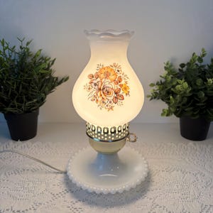 Peut inclure: Une lampe de table blanche vintage avec un motif floral. La lampe a un abat-jour en verre dépoli avec un bord festonné et une base avec un bord festonné. Le motif floral comprend des fleurs orange, jaunes et marron. La lampe est branchée et allumée.