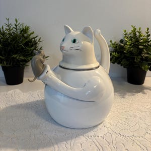 Może przedstawiać: Biały ceramiczny czajnik w kształcie kota, trzymający małą szarą mysz. Czajnik ma zielone oczy i czarne detale. Uchwyt jest zakrzywiony, a dzióbek znajduje się na piersi kota. Na białym koronkowej obrusie.