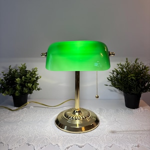 May include: A banker's lamp featuring a polished gold-tone metal base and a cylindrical green glass shade. The lamp includes a vertical stand, a circular tiered base, and a gold pull chain with a small spherical weight for turning the light on.