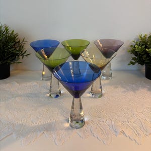Peut inclure: Un ensemble de six verres à martini avec des bols colorés dans des tons de bleu, vert et violet, chacun doté d'un pied et d'une base en verre transparent, épais et conique. Les verres sont disposés en groupe, révélant leurs teintes vives.