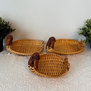 May include: Three duck-shaped woven baskets. The oval baskets have a woven pattern and duck heads at each end, made of a darker brown material. They are displayed on a white lace tablecloth, with green plants in the background.