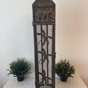 May include: A tall, bronze-colored metal decorative cabinet with a mesh front and a peaked roof. The top features an embossed elephant design. Palm tree cutouts adorn the front. The cabinet is flanked by two small potted green plants.