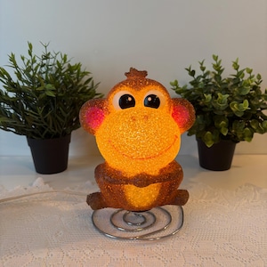 Puede incluir: Una figura de mono iluminada con un cuerpo naranja brillante, detalles marrones y ojos negros grandes. El mono está sentado sobre una base de metal plateado y tiene orejas rosas. Dos pequeñas plantas en macetas están a cada lado del mono.