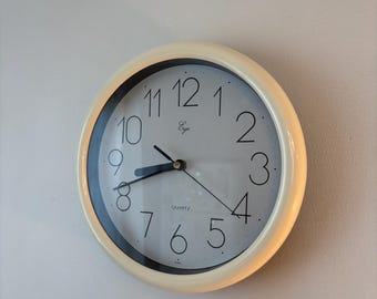 Puede incluir: Un reloj de pared redondo con un marco de color crema y una esfera blanca. El reloj presenta números arábigos negros del 1 al 12, manecillas negras para las horas y los minutos, un segundero negro fino y el texto "Ergo" y "QUARTZ" impresos.