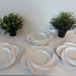 Peut inclure: Ensemble d'assiettes blanches en forme de pétales disposées sur une surface blanche. Deux petites plantes vertes en pot encadrent les assiettes. Les assiettes présentent un design tourbillonnant distinctif, idéal pour servir des entrées ou des desserts.