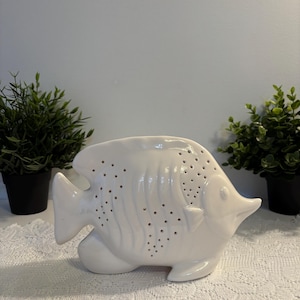 Peut inclure: Un objet décoratif en céramique blanche en forme de poisson, percé de nombreux petits trous. Le poisson a un corps incurvé, des nageoires et un museau pointu. Il est posé sur une nappe en dentelle blanche, avec des plantes en pot en arrière-plan.