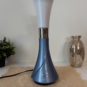 Peut inclure: Lampe de bureau OttLite bleue et argentée avec une base évasée et un abat-jour blanc en forme de cône. La lampe a une bande argentée et un bouton d'alimentation. Un cordon d'alimentation noir part de la base. Une petite plante en pot et un vase décoratif sont en arrière-plan.