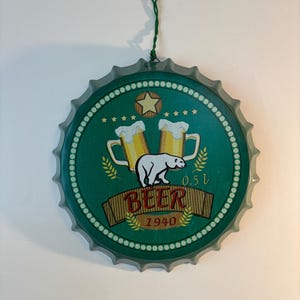 May include: A circular metal wall sign shaped like a bottle cap with a green background. It features a white polar bear, two beer mugs with foam, a star, and wheat stalks. The text reads BEER and 1940, with a 0.5 l capacity label.