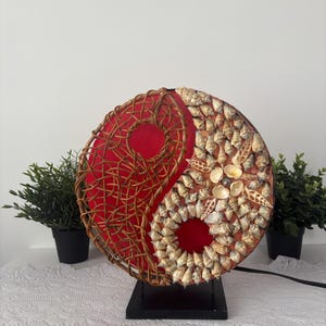 Peut inclure: Lampe décorative Yin Yang avec un fond en tissu rouge. Un côté est tissé avec un matériau marron, l'autre est orné de coquillages. La lampe est montée sur une base noire et possède un cordon d'alimentation.