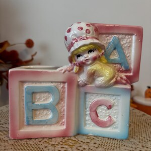 Peut inclure: Un pot de fleurs vintage en céramique en forme de blocs d'alphabet, avec les lettres "B", "C" et "A". Une petite figurine d'une petite fille portant un bonnet regarde par-dessus les blocs. Les blocs sont peints en rose pastel et bleu.