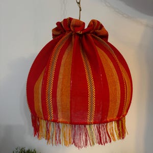 Peut inclure: Abat-jour en tissu ray&eacute; rouge et orange avec une frange en bas. L'abat-jour est suspendu &agrave; une cha&icirc;ne dor&eacute;e. Le tissu pr&eacute;sente des rayures verticales rouges et oranges avec un haut fronc&eacute;.