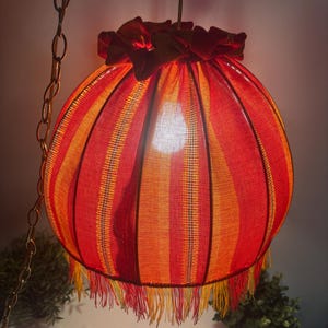 Peut inclure: Une lampe suspendue vintage avec un abat-jour sph&eacute;rique. L'abat-jour pr&eacute;sente des rayures verticales rouges, oranges et jaunes. La lampe est orn&eacute;e d'une frange et d'un volant sur le dessus. Elle est suspendue &agrave; une cha&icirc;ne en m&eacute;tal.