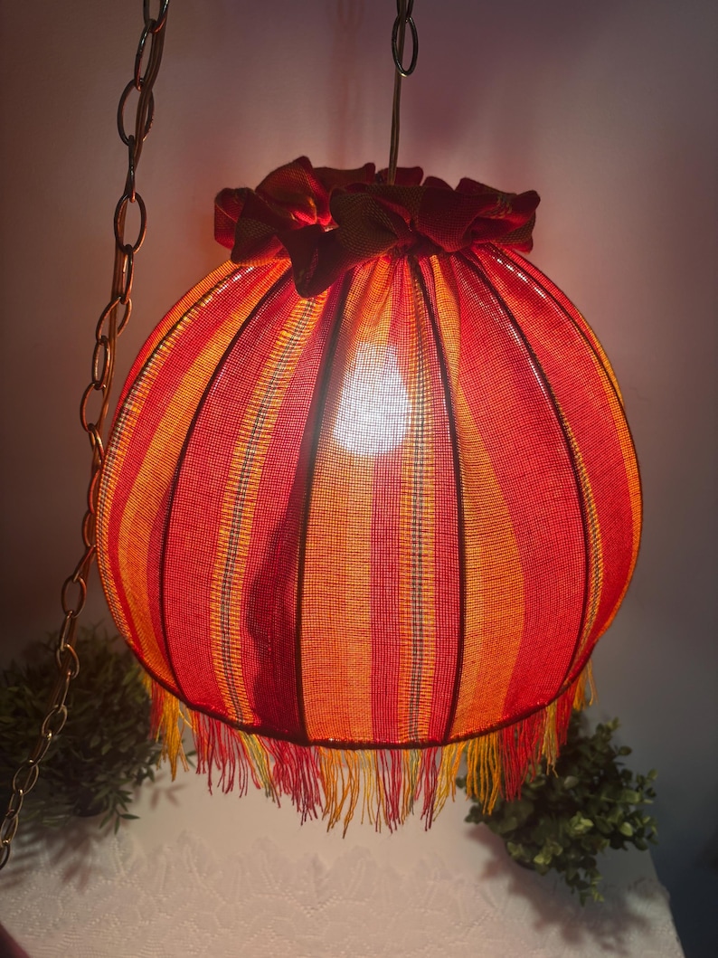 Peut inclure: Une lampe suspendue vintage avec un abat-jour sph&eacute;rique. L'abat-jour pr&eacute;sente des rayures verticales rouges, oranges et jaunes. La lampe est orn&eacute;e d'une frange et d'un volant sur le dessus. Elle est suspendue &agrave; une cha&icirc;ne en m&eacute;tal.