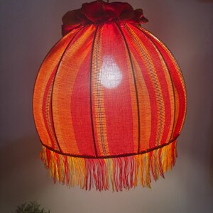 Peut inclure: Suspension luminaire avec abat-jour rond ray&eacute; rouge et orange. L'abat-jour est orn&eacute; d'une frange en bas et est suspendu &agrave; une cha&icirc;ne m&eacute;tallique. La lampe est &eacute;clair&eacute;e de l'int&eacute;rieur.