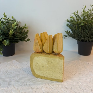 May include: A wooden cheese sculpture featuring a cheese wedge and several cheese wedges on top. The cheese is painted in shades of yellow and tan. The sculpture is on a white surface. Green plants are in the background.