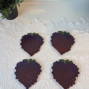 Peut inclure: Quatre dessous de verre en forme de grappe de raisin, avec une surface texturée violet foncé et des accents verts. Les dessous de verre sont disposés sur une nappe en dentelle blanche, avec une petite plante en pot en arrière-plan.