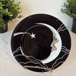May include: A black decorative plate featuring a crescent moon with a face, a shooting star, and a star. The plate has a white and orange face, with white clouds and a white border. The plate is displayed on a gold-colored stand.
