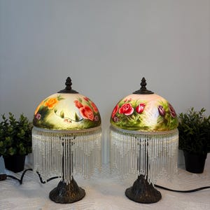 Può includere: Una coppia di lampade da tavolo con paralumi in vetro a cupola decorati con rose dipinte a mano in rosso, arancione e viola su sfondo verde. Ogni lampada ha una base in metallo scuro e una lunga frangia di perline trasparenti sul bordo.