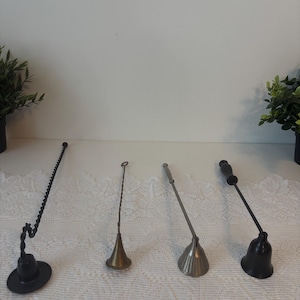 May include: Four candle snuffers of varying designs are arranged on a white lace tablecloth. The snuffers are made of metal and feature shapes like a bell and a cone. The colors include black, silver, and brass. The items are for use with candles.