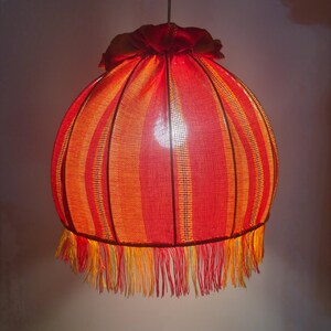 Peut inclure: Suspension luminaire sph&eacute;rique avec abat-jour ray&eacute; rouge et orange. L'abat-jour est orn&eacute; d'une frange et d'un dessus en tissu fronc&eacute;. La lampe est suspendue &agrave; une cha&icirc;ne m&eacute;tallique. Diam&egrave;tre d'environ 30 cm.