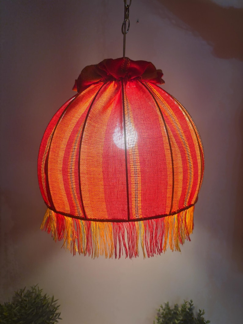 Peut inclure: Suspension luminaire sph&eacute;rique avec abat-jour ray&eacute; rouge et orange. L'abat-jour est orn&eacute; d'une frange et d'un dessus en tissu fronc&eacute;. La lampe est suspendue &agrave; une cha&icirc;ne m&eacute;tallique. Diam&egrave;tre d'environ 30 cm.