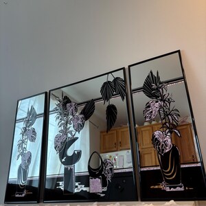 May include: A set of three rectangular wall mirrors with black frames, each featuring a decorative black and glittery pink floral design. The artwork depicts stylized potted plants and vases, with reflective surfaces that show the surrounding room.