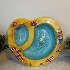 Puede incluir: Una fuente de cerámica en forma de corazón con dos piscinas azules y un borde amarillo. La fuente presenta ilustraciones de personas y objetos asociados con el verano, incluyendo una sirena, una radio y una persona leyendo las noticias.