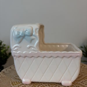 May include: A white ceramic baby cradle planter with a light blue bow and pink trim. The planter features a diamond pattern and sits on four small feet. A decorative item for nurseries or baby showers.