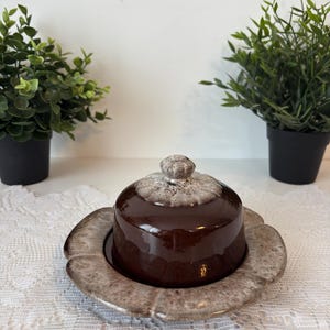 Peut inclure: Un beurrier en céramique marron avec un couvercle, posé sur une assiette assortie. Le plat a une finition brillante et une base texturée en forme de fleur. Le couvercle a un petit bouton texturé. Deux petites plantes en pot sont en arrière-plan.