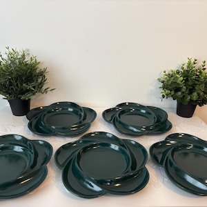 Peut inclure: Six assiettes en céramique vert foncé avec un design tourbillonnant unique sont disposées sur une surface blanche. Les assiettes ont la forme d'une fleur. Deux petites plantes en pot sont de chaque côté des assiettes.