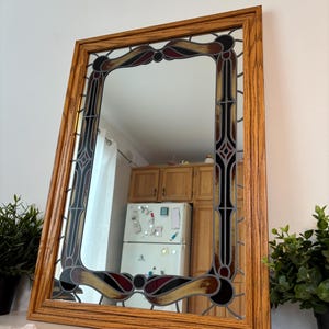 Peut inclure: Miroir rectangulaire avec un motif de vitrail et un cadre en bois. Le vitrail présente des accents noirs, rouges et dorés. Le miroir est incliné, reflétant une pièce avec des meubles en bois et un réfrigérateur.