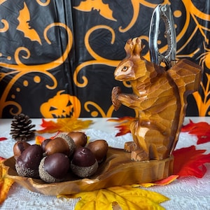 May include: A wooden squirrel nutcracker with a silver nut-cracking tool. The squirrel holds an acorn and sits on a wooden base with a small wooden dish filled with acorns. The background includes fall leaves and a Halloween-themed backdrop.