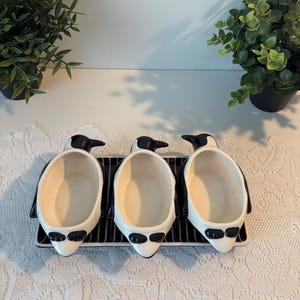 Peut inclure: Un ensemble de trois bols en céramique en forme de pingouin disposés sur un plateau rectangulaire à rayures noires et blanches. Chaque bol présente un extérieur noir et blanc avec un intérieur crème lisse, imitant la forme d'un pingouin.