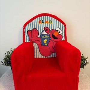 May include: A red children's armchair featuring Elmo from Sesame Street. The chair has the name "ELMO" on the backrest, along with an image of Elmo reading a book. The chair is designed for young children and has a soft, furry texture.