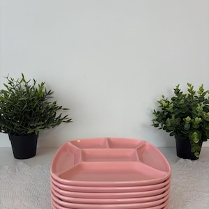 Peut inclure: Une pile de huit assiettes roses divisées. Chaque assiette comporte quatre compartiments. Les assiettes sont empilées sur une nappe en dentelle blanche. Deux petites plantes vertes en pot sont de chaque côté des assiettes.