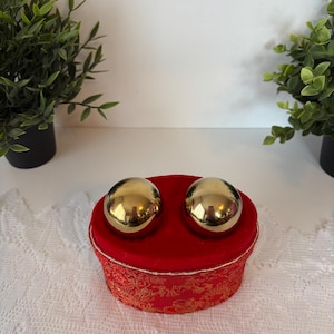 Puede incluir: Dos esferas doradas descansan sobre una caja redonda roja. La caja tiene una parte superior de terciopelo rojo y un patrón dorado en los lados. Las esferas reflejan el entorno. La caja está sobre una superficie blanca.