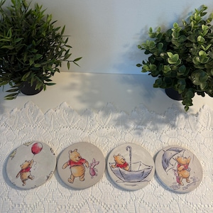 May include: A set of four round coasters featuring Winnie the Pooh illustrations. Each coaster depicts a different scene, including Pooh with a balloon, Piglet, and an umbrella. The coasters are beige with a white border.