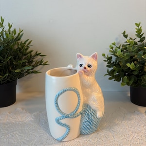 Op de afbeelding: Een cilindrische vaas van wit keramiek met een gebeeldhouwd wit kitten aan de zijkant. Het kitten rust met zijn pootjes op een blauwe bol wol met textuur die overgaat in een blauwe touwstreng die om de voorkant van de vaas heen loopt.