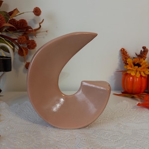 Peut inclure: Une sculpture en céramique pêche en forme de croissant de lune. La surface lisse et brillante reflète la lumière, soulignant la forme incurvée. La pièce décorative repose sur une nappe en dentelle blanche, avec des décorations sur le thème de l'automne en arrière-plan.