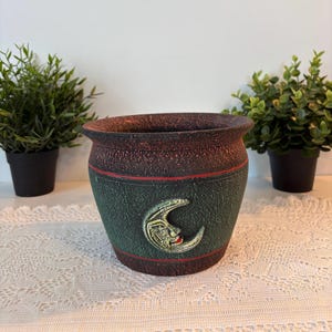 Peut inclure: Un pot de fleurs rond et texturé avec un bord supérieur brun foncé et un corps vert forêt. Un visage de croissant de lune sculpté en relief avec des lèvres rouges est centré sur le devant. Le pot présente de fines rayures rouges horizontales.
