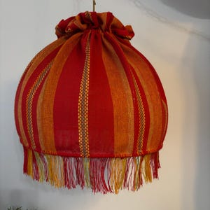 Peut inclure: Abat-jour suspendu avec un motif &agrave; rayures rouges et oranges. L'abat-jour a une forme ronde avec un dessus fronc&eacute; et une bordure &agrave; franges en bas. La frange comprend des fils rouges, jaunes et verts. L'abat-jour est suspendu par une cha&icirc;ne dor&eacute;e.